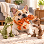 Tan French Bulldog holding an orange plush dinosaur dog toy on a cream carpet, with green and light brown dinosaur toys nearby in a bright living room.